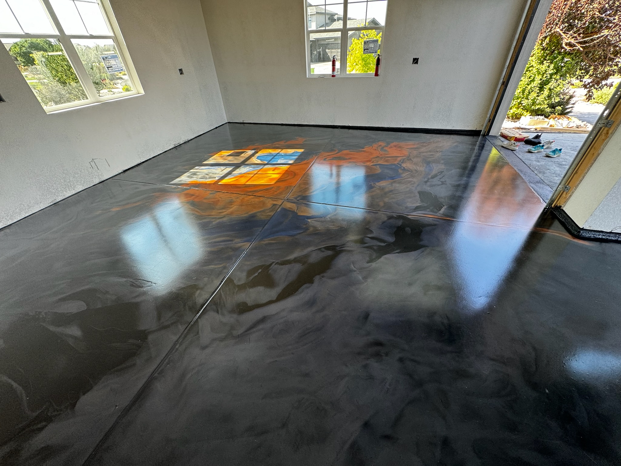 Black metallic epoxy garage floor with orange and blue veining, a window's reflection on the gloss and the overhead door open to daylight